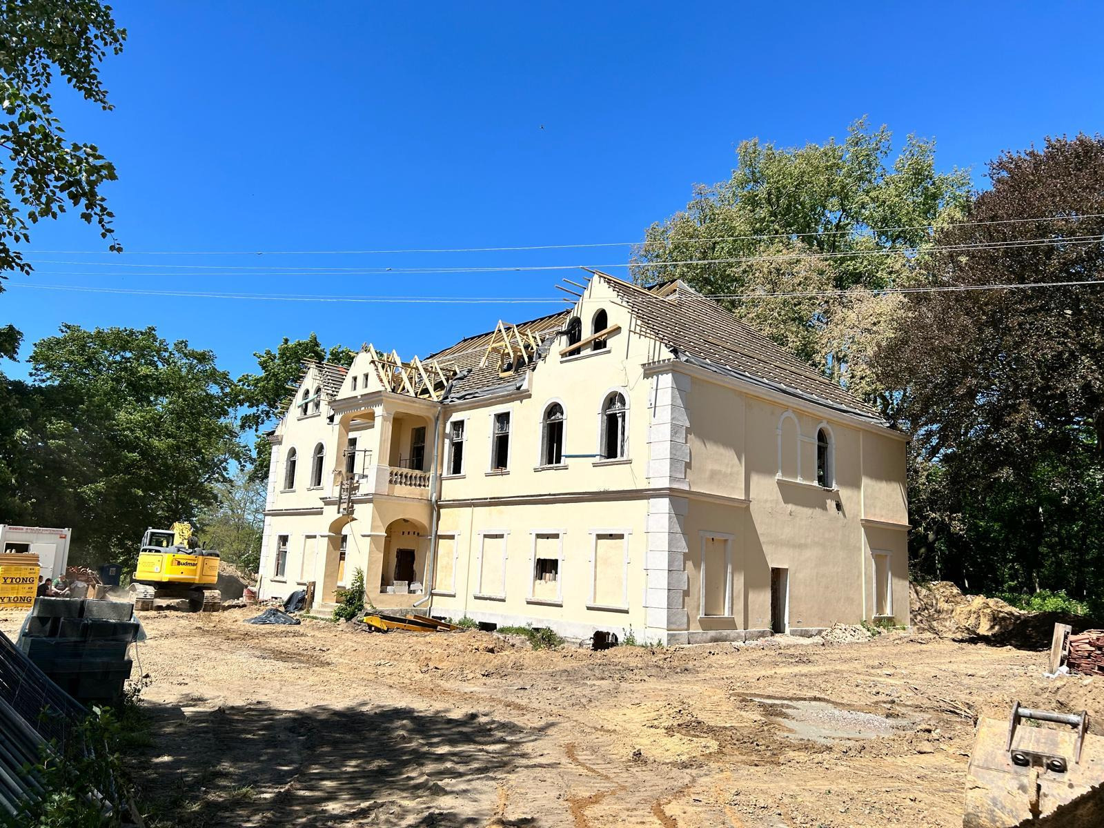 Remontowany, dwukondygnacyjny budynek z kolumnami i balkonem, widoczny szkielet dachu, koparka w tle, teren budowy.