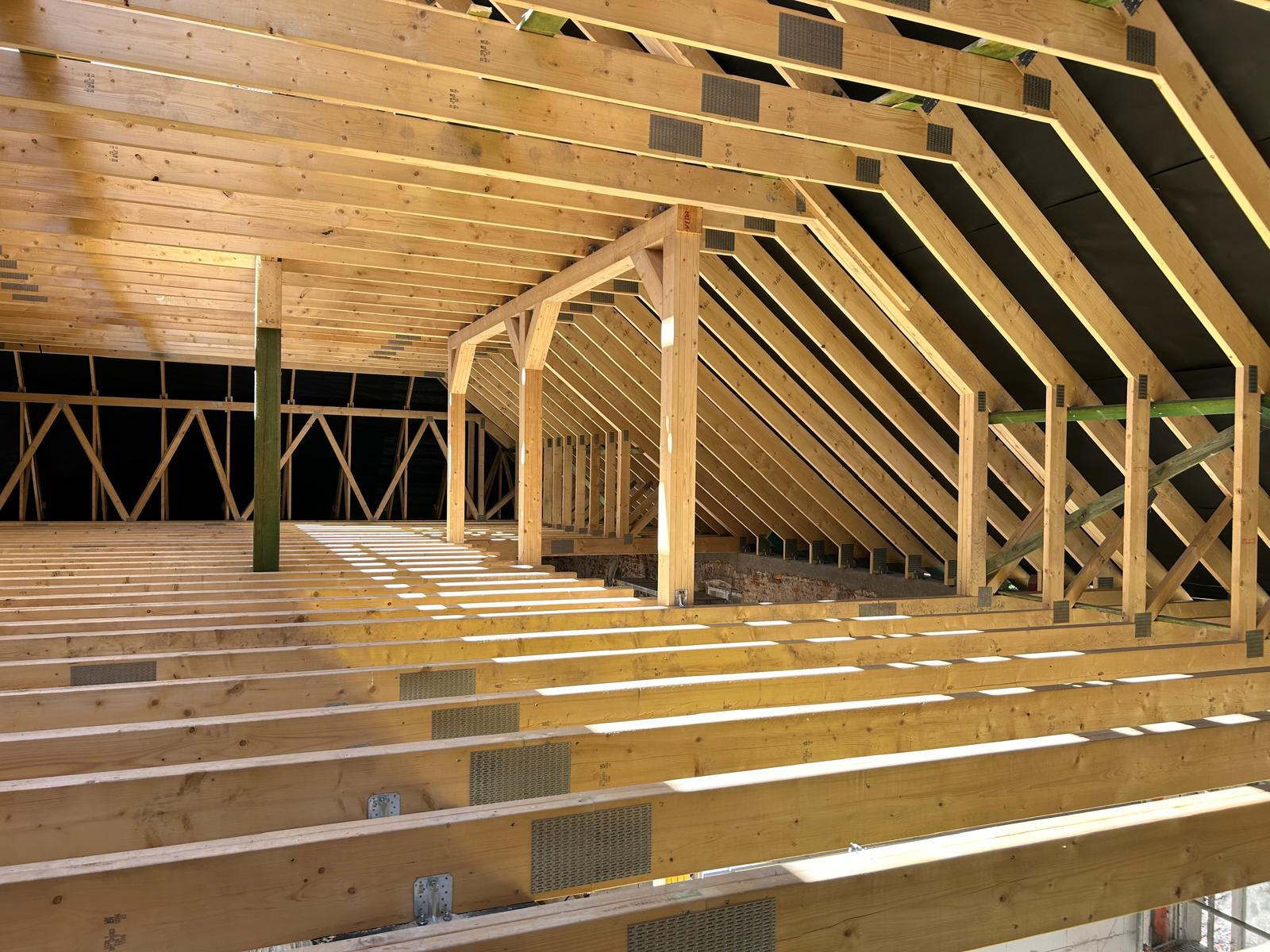 Wnętrze w trakcie budowy: drewniana konstrukcja szkieletowa dachu i podłogi, widoczne elementy mocujące i membrany izolacyjne, oświetlone promieniami słonecznymi wpadającymi przez otwory.