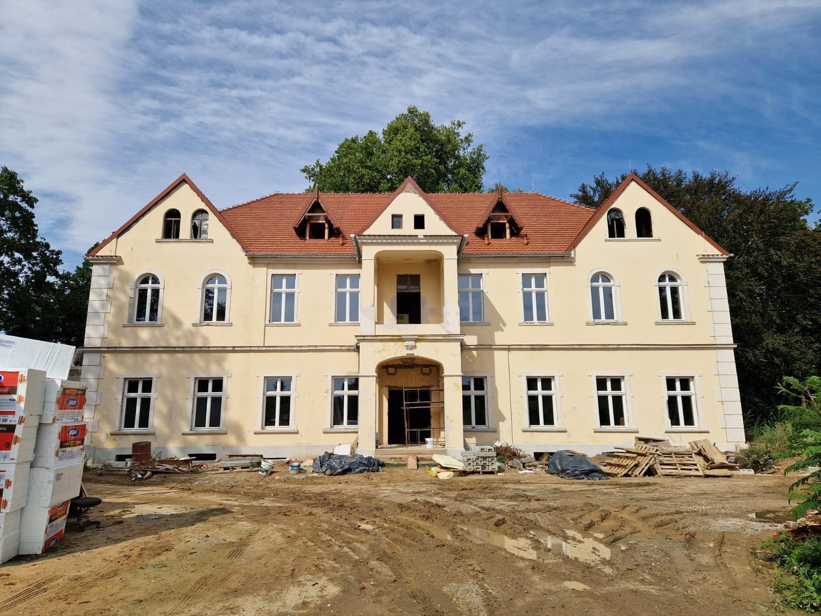 Żółty, dwupiętrowy budynek z czerwoną dachówką w trakcie renowacji; widoczne rusztowania w wejściu, materiały budowlane i błotniste podłoże.