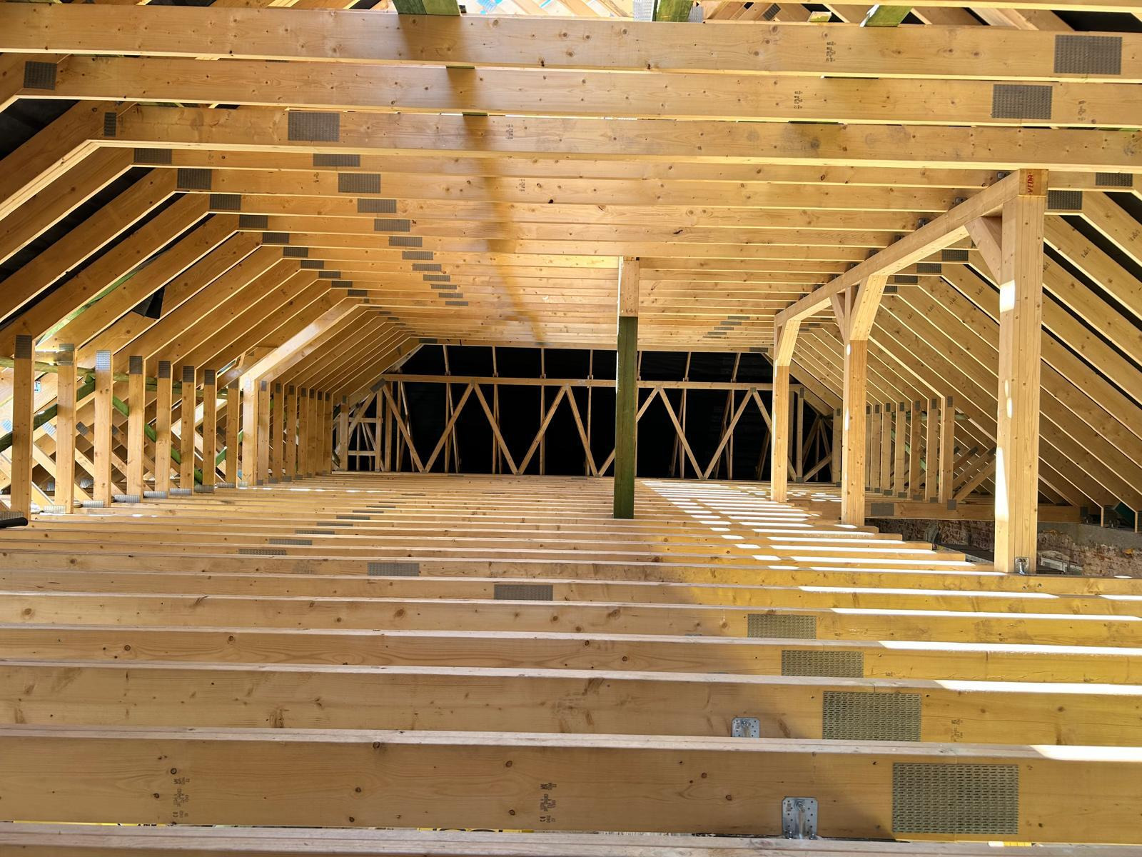 Wnętrze w trakcie budowy z drewnianą konstrukcją dachu i podłogi, widoczne metalowe łączniki i ciemne tło w oddali.