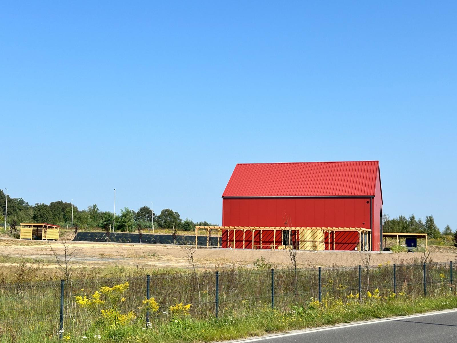 Nowoczesny, czerwony budynek z blachy trapezowej z drewnianą konstrukcją pergoli w stanie surowym, na tle błękitnego nieba i przygotowanego terenu.