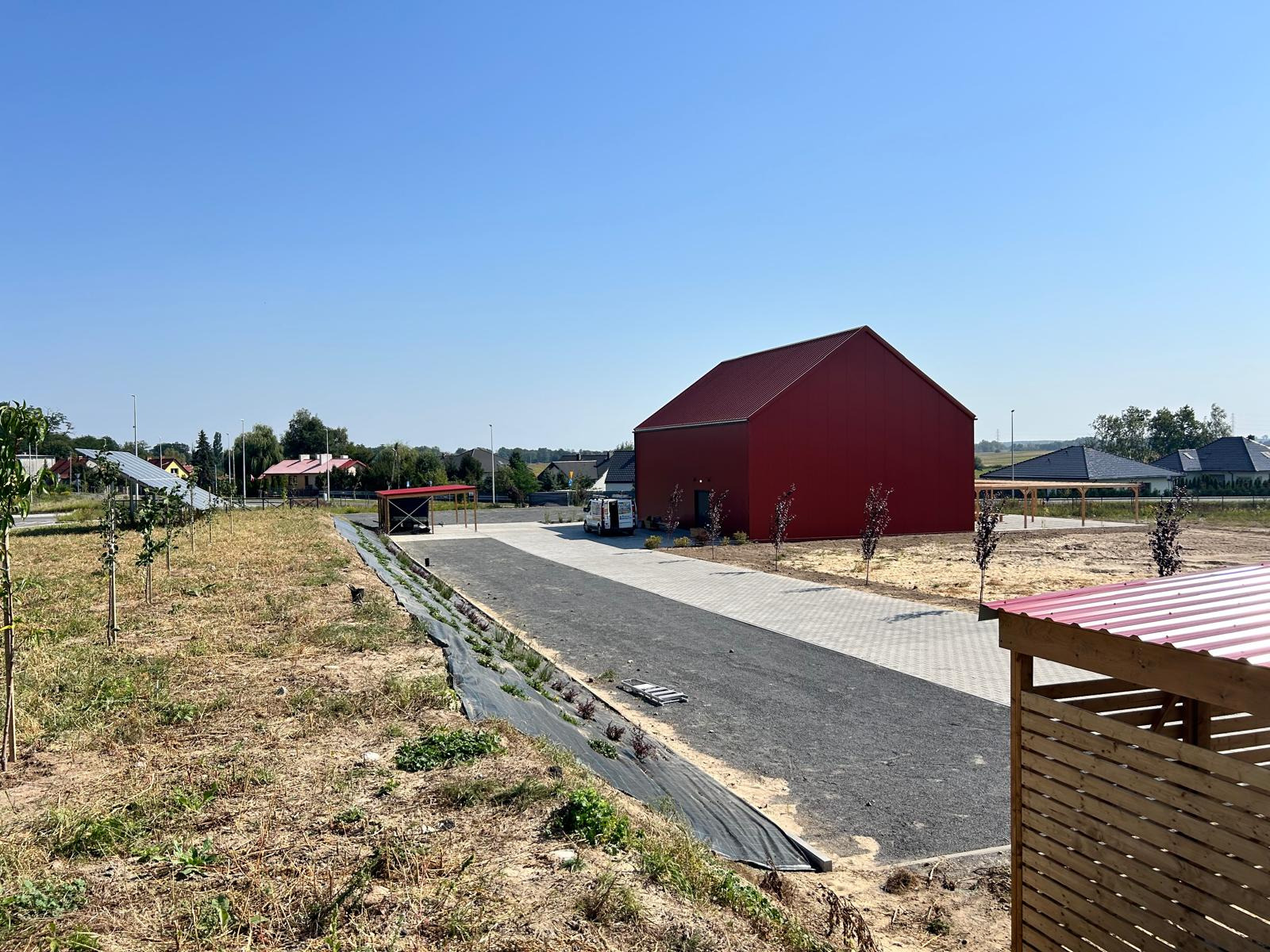 Nowoczesny, czerwony budynek gospodarczy z dwuspadowym dachem, widok z zewnątrz na tle błękitnego nieba i zabudowań mieszkalnych w oddali, z utwardzonym terenem i młodymi drzewkami.