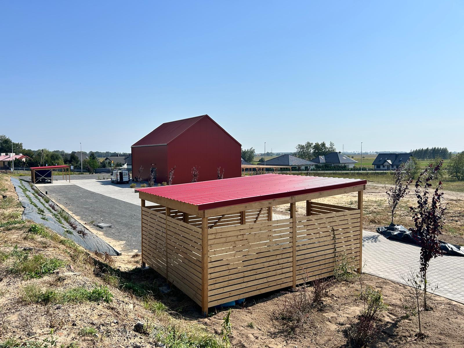 Nowo wybudowana drewniana wiata z czerwonym dachem na tle czerwonego budynku i domów jednorodzinnych, świeżo zasadzone drzewka w pobliżu.