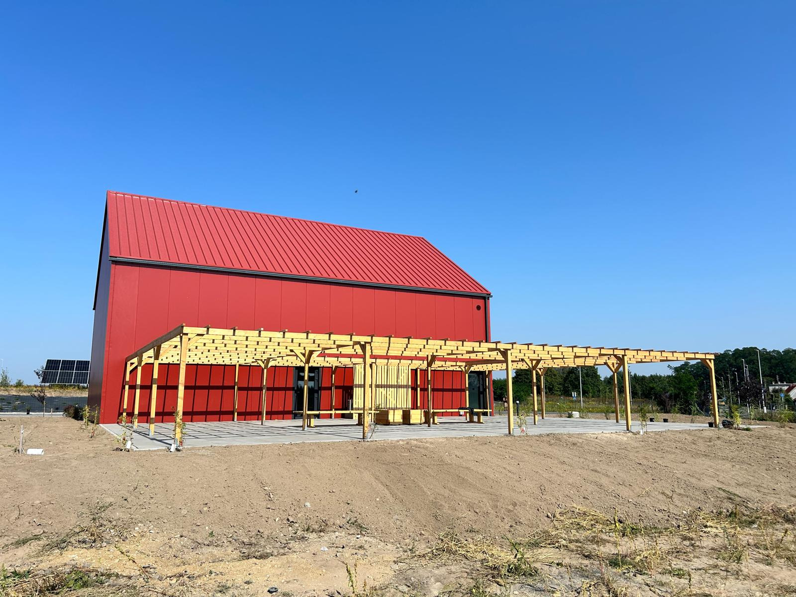 Nowoczesny budynek o czerwonej fasadzie i dachu z blachy, z drewnianą pergolą na betonowym fundamencie, na tle błękitnego nieba.
