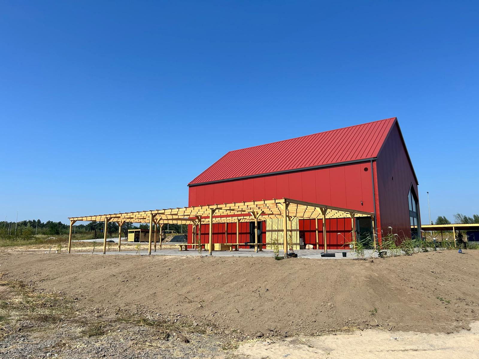 Nowoczesny, czerwony budynek z metalowym dachem i drewnianą pergolą w trakcie budowy, widok z poziomu gruntu na tle błękitnego nieba.