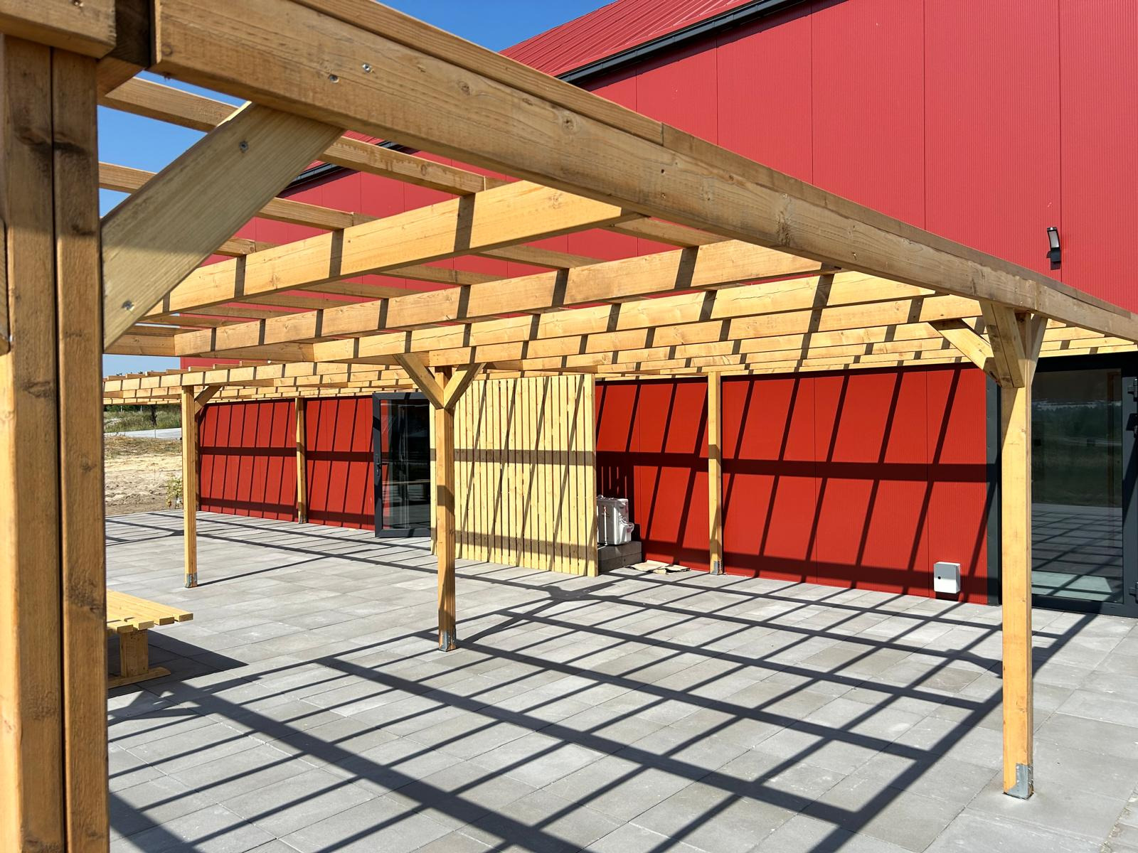Drewniana pergola w surowym stanie, rzucająca geometryczny cień na szarą kostkę brukową, na tle czerwonej ściany budynku z ryflowanego metalu i fragmentu żółtej drewnianej ściany.
