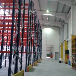 Wnętrze hali magazynowej z zamontowanymi regałami wysokiego składowania w kolorach czarnym, srebrnym i czerwonym, zabezpieczonymi żółtymi odbojnicami, oświetlone lampami przemysłowymi.