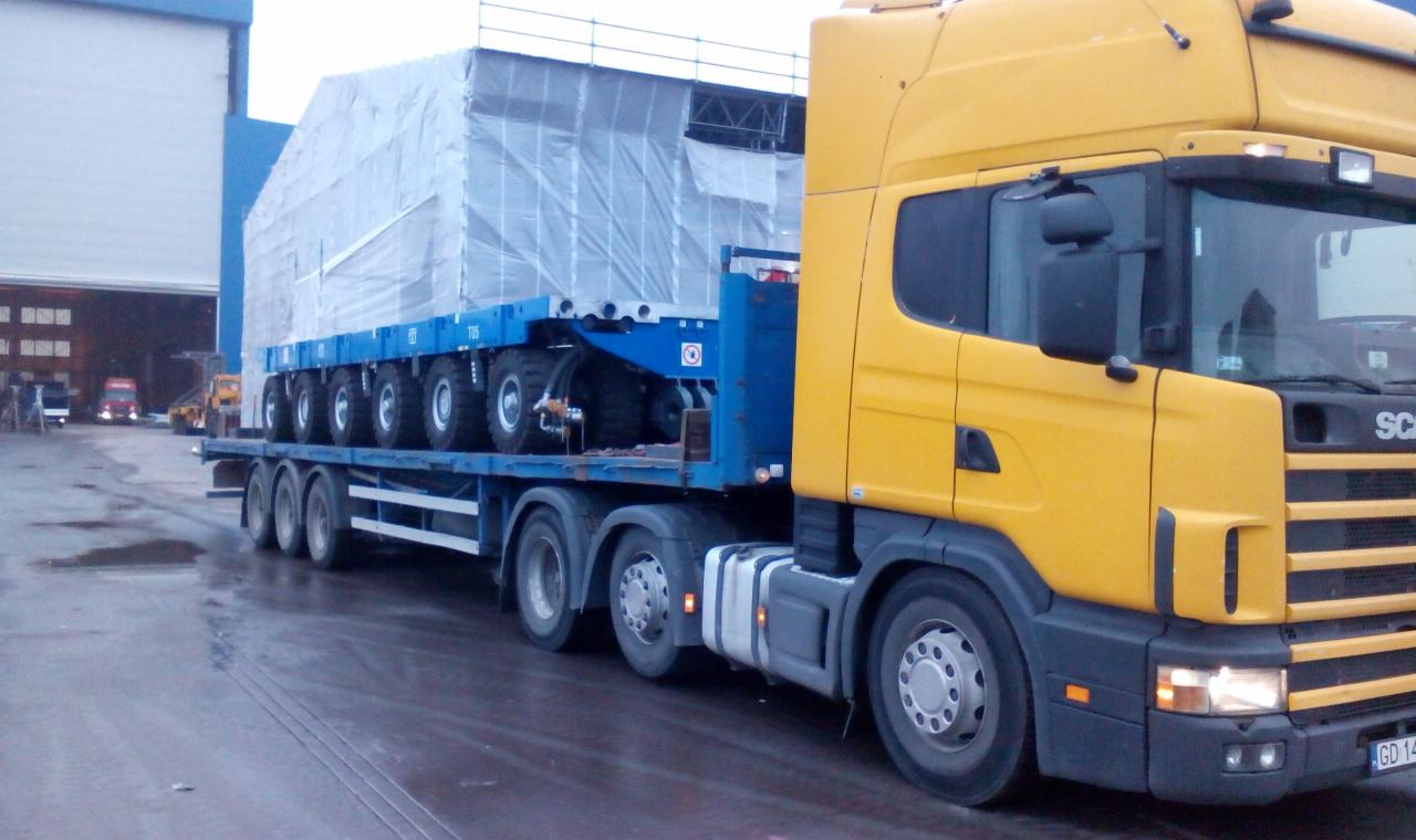 Żółta ciężarówka Scania przewozi na niebieskiej, wieloosiowej platformie ładunek owinięty srebrną folią, widoczne tablice rejestracyjne GD 14.