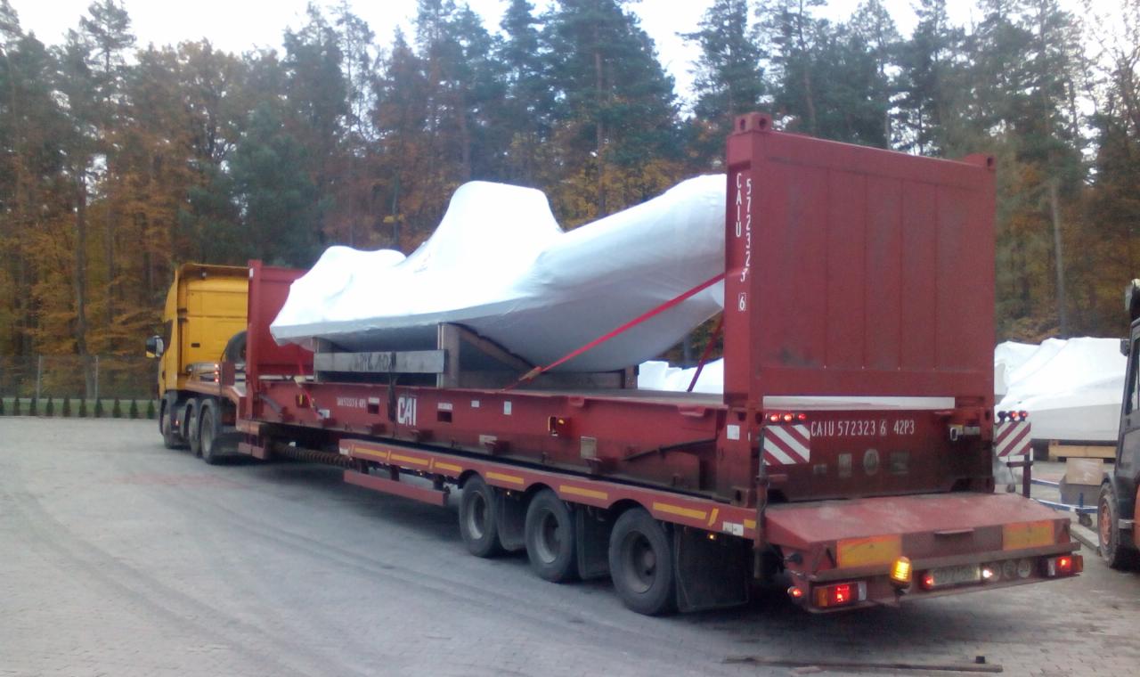 Żółta ciężarówka z czerwoną platformą przewozi biały, owinięty ładunek o nieregularnym kształcie, zabezpieczony czerwonymi pasami, na tle jesiennego lasu.