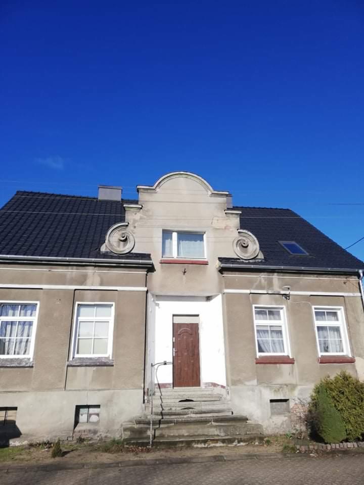 Dwukondygnacyjny dom z ciemnym, skośnym dachem i ozdobnymi elementami nad wejściem, sfotografowany w słoneczny dzień.