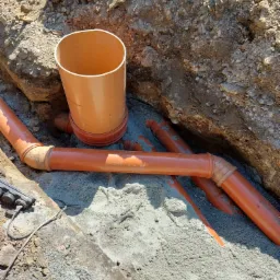 Pomarańczowe rury kanalizacyjne ułożone w wykopie ziemnym, częściowo zasypane piaskiem, z widocznym złączem kolankowym.