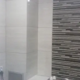 Ściana wyłożona jasnymi, kwadratowymi płytkami ceramicznymi oraz fragment ściany z dekoracyjnymi, poziomymi paskami płytek w odcieniach szarości i czerni, widoczne narzędzia na blacie.