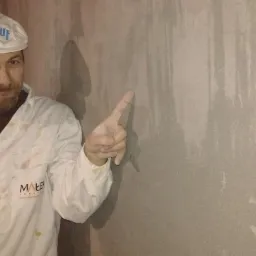 Tynkarz w ubraniu roboczym marki KNAUF wskazuje palcem na gładką, szarą ścianę po tynkowaniu. Widoczne ślady po aplikacji maszynowej.