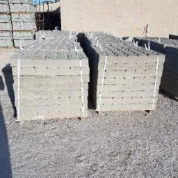 Stosy szarych płyt betonowych ułożonych na paletach, zabezpieczonych białym sznurkiem, na żwirowym podłożu, z cieniem osoby widocznym po lewej stronie.