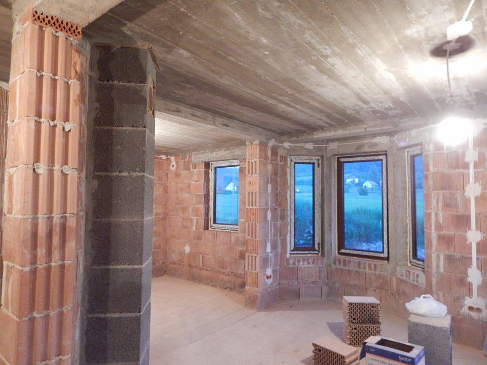 Wnętrze w stanie surowym: murowane ściany z czerwonej cegły, betonowy strop, okna z widokiem na zewnątrz, instalacja elektryczna w peszlach na ścianie, materiały budowlane na podłodze.