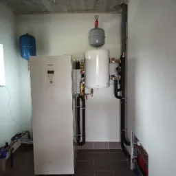 Pompa ciepła Panasonic Aquarea, zbiornik buforowy i naczynia wzbiorcze w kotłowni z surowym betonowym stropem i rozdzielaczem ogrzewania podłogowego w ścianie.