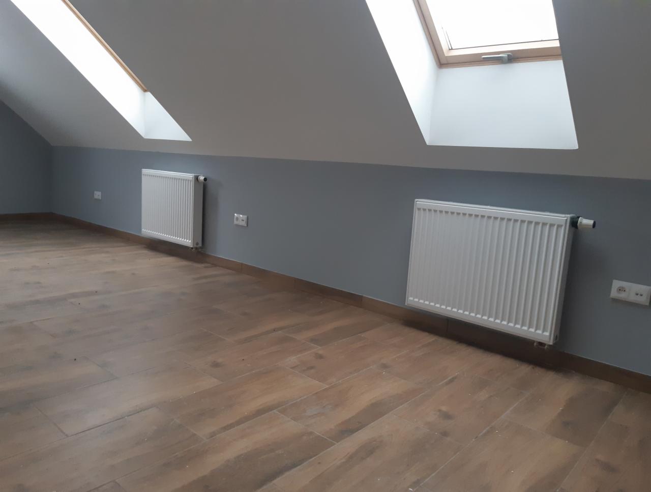 Poddasze z dwoma białymi grzejnikami panelowymi pod oknami dachowymi, szare ściany i podłoga imitująca drewno.