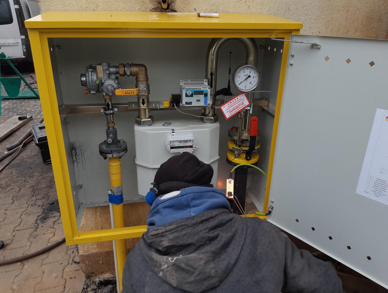 Montera w trakcie podłączania gazomierza w żółtej szafce, widoczny manometr, zawory i instalacja gazowa.