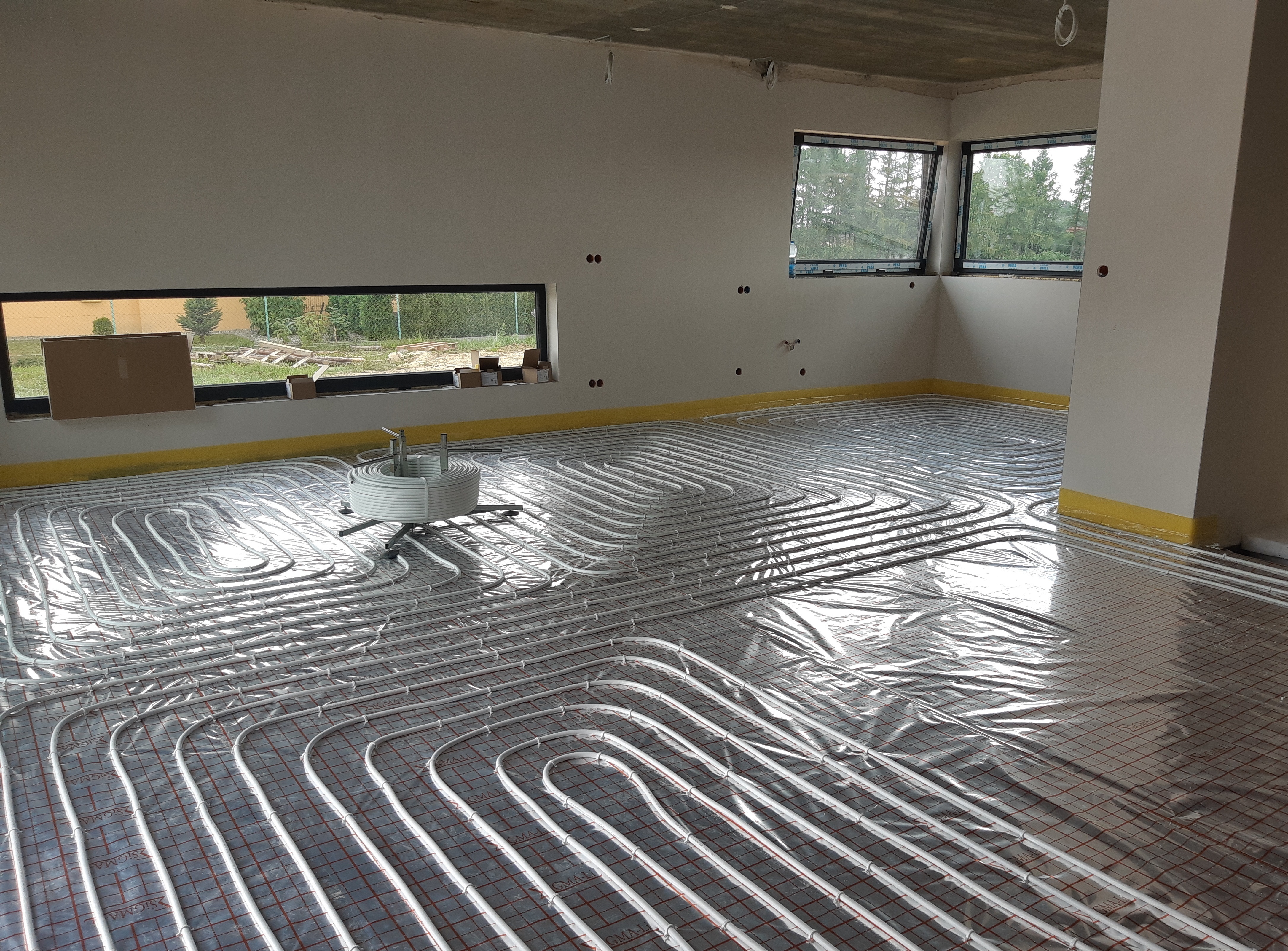 Rozłożone rury ogrzewania podłogowego na folii izolacyjnej z widocznym rozdzielaczem w surowym wnętrzu z oknami.