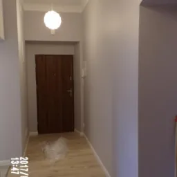 Pomieszczenie po malowaniu ścian na szaro, widoczna drewniana podłoga, drzwi wejściowe i lampy sufitowe, na podłodze leżą resztki folii malarskiej, w rogu stoi miotła.