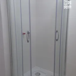 Narożna kabina prysznicowa z przesuwnymi drzwiami, zamontowana w łazience wyłożonej białymi płytkami, widoczne narzędzia i folia ochronna na podłodze.