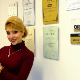 Kobieta w bordowym golfie prezentuje złotą statuetkę na tle ściany z dyplomami i nagrodami, w tym tabliczką 'Orły Prawa 2018 - Laureat Konkursu Wybór Klientów'.