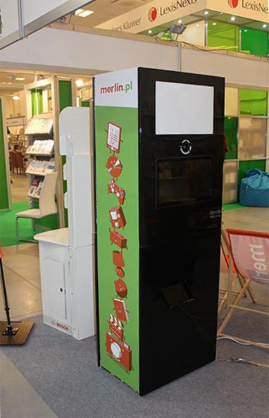 Automat do zdjęć z ekranem, panelem dotykowym i grafiką na zielonym tle z logiem merlin.pl, ustawiony na szarym dywanie targowym obok stoiska z ekspozycją produktów.