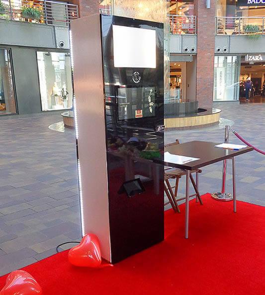 Automat do zdjęć z ekranem i panelem dotykowym, ustawiony na czerwonym dywanie w galerii handlowej, obok stolik i krzesło, dekoracja w postaci czerwonych balonów w kształcie serca.