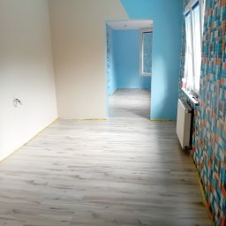 MarBud - Wnętrze po odświeżeniu: jasne panele podłogowe, ściany częściowo pomalowane na biało i błękitno, z akcentem w postaci wzorzystej tapety na jednej ścianie, oklejone taśmą malarską przy podłodze.