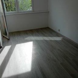 MarBud - Jasne panele podłogowe ułożone w pustym pokoju z białymi ścianami, oświetlone naturalnym światłem wpadającym przez okno.