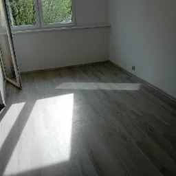 Jasne panele podłogowe ułożone w pustym pokoju z białymi ścianami, oświetlone naturalnym światłem wpadającym przez okno.