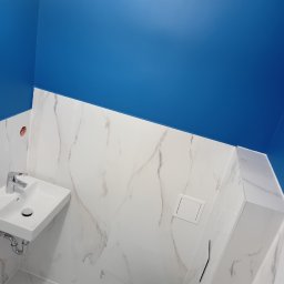 MarBud - Wykończona łazienka z niebieską ścianą, białymi marmurkowymi płytkami, wiszącą umywalką i podwieszanym WC z zabudową.