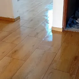 Połyskująca, świeżo położona podłoga z płytek imitujących drewno w korytarzu, widoczne łączenia płytek i listwy przypodłogowe.