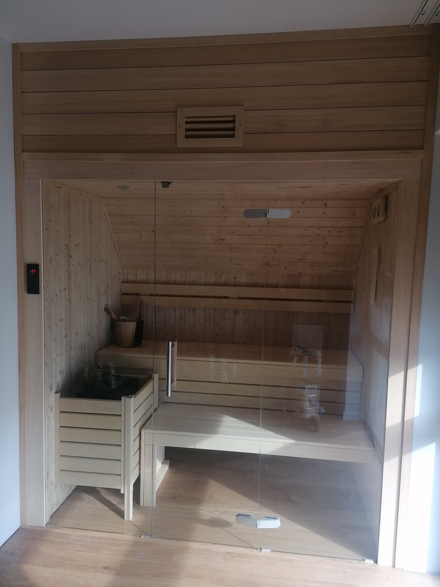 Nowoczesna sauna z jasnego drewna, z przeszklonymi drzwiami i cyfrowym termometrem na ścianie, ukazująca drewniane ławki, piec z kamieniami i akcesoria do sauny.