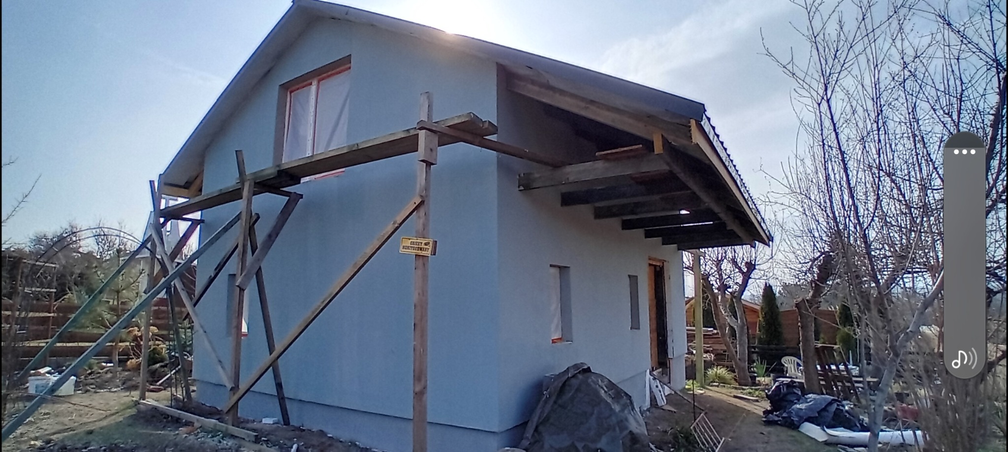 Dom jednorodzinny w trakcie prac wykończeniowych elewacji, widoczne drewniane rusztowania, okno zabezpieczone folią, fragment zadaszenia werandy w trakcie budowy.