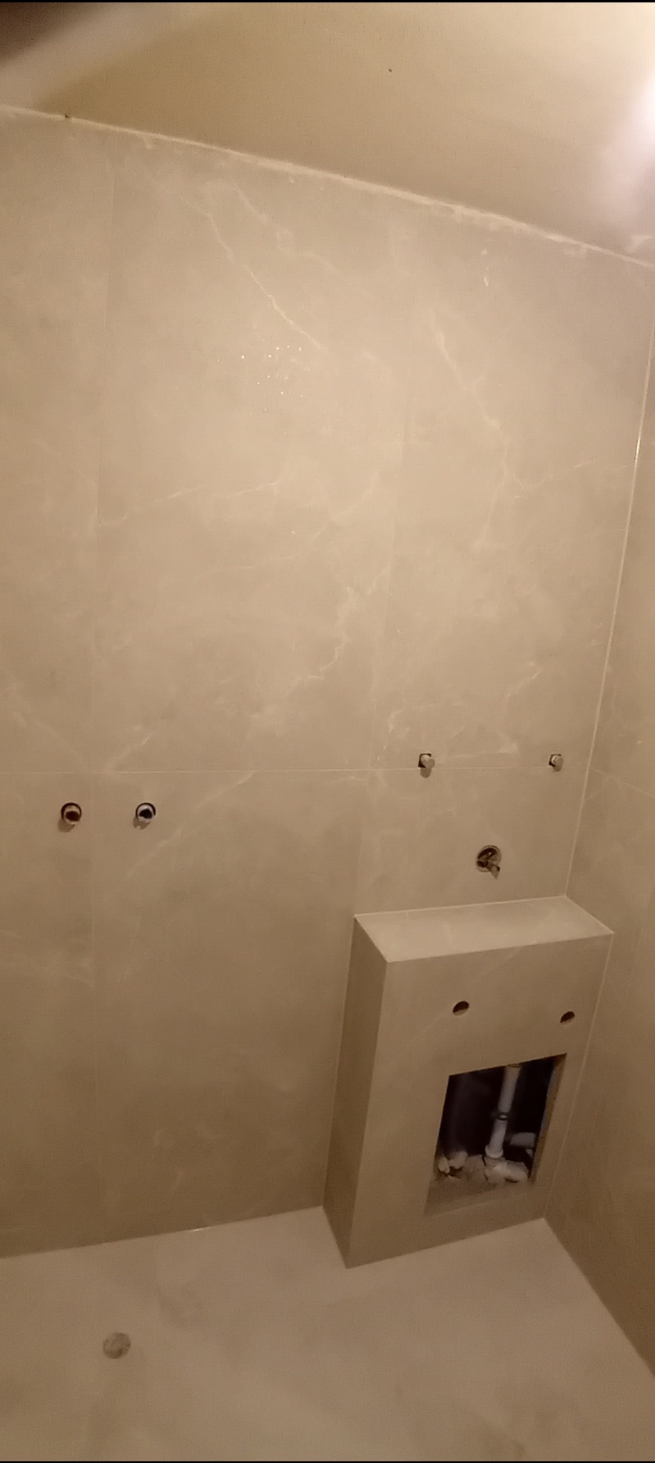 Ściana łazienki wyłożona jasnymi płytkami imitującymi marmur, z przygotowanymi punktami instalacyjnymi i zabudową geberitu w trakcie prac instalacyjnych.