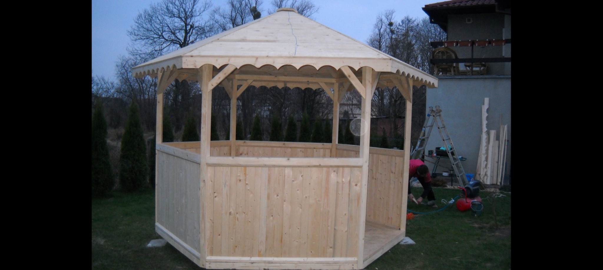Drewniana, surowa altana w trakcie budowy, widoczne narzędzia i osoba pracująca wewnątrz konstrukcji, na trawniku, w tle drzewa i budynek.