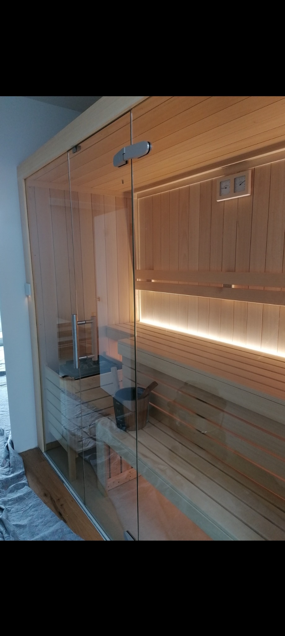 Nowoczesna sauna z przeszklonymi drzwiami, drewnianymi ławkami i oświetleniem LED w listwach, wiaderko z chochlą w rogu, termometry na ścianie, podłoga częściowo zakryta folią ochronną.