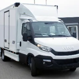 Biały samochód dostawczy Iveco z zabudową kontenerową, zaparkowany na brukowanej powierzchni przed budynkiem.