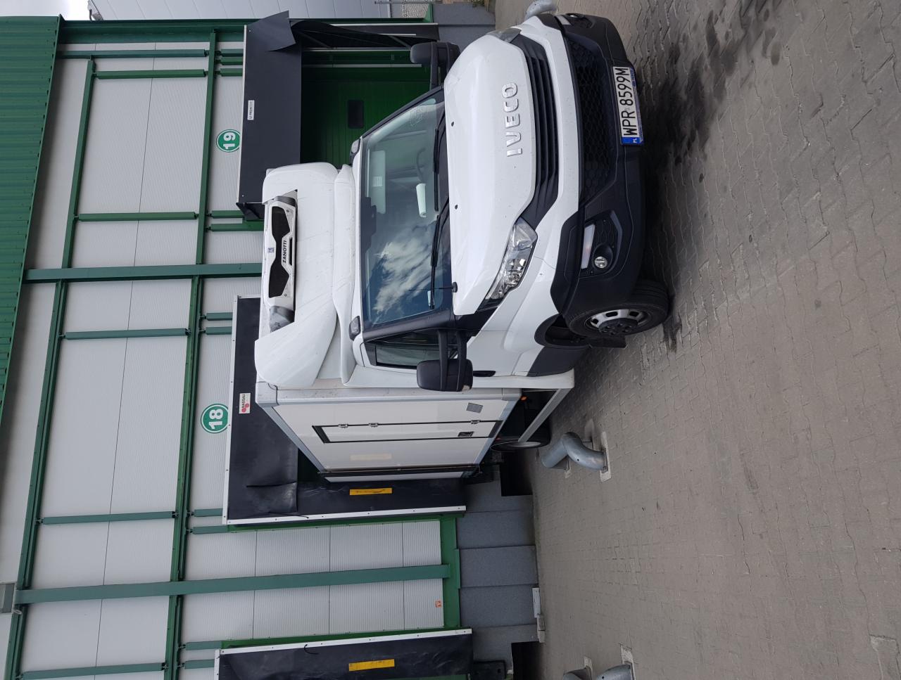 Biały, chłodniczy samochód dostawczy Iveco z numerem rejestracyjnym WPR 8599M, zaparkowany przy rampie załadunkowej magazynu z numerem 18 i 19.