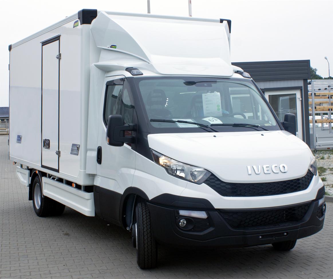 Biały samochód dostawczy Iveco z zabudową kontenerową, zaparkowany na brukowanej powierzchni przed budynkiem.