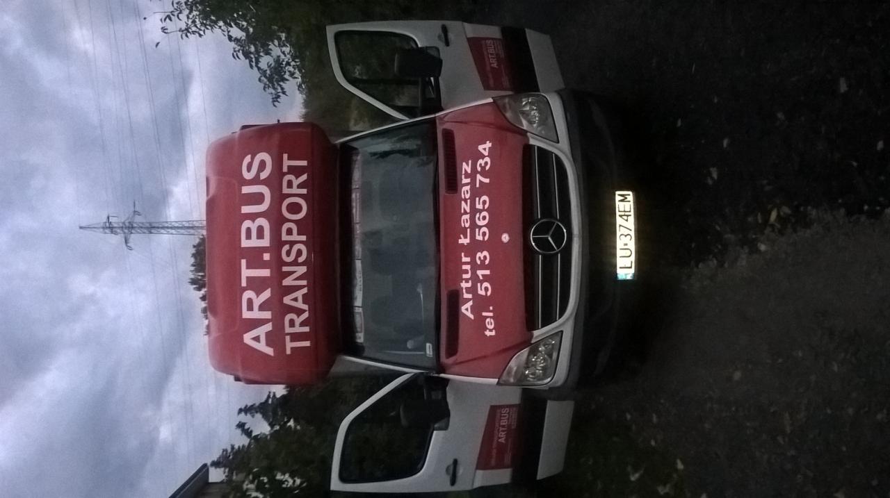 Biało-czerwony bus transportowy z otwartymi drzwiami, widoczne dane kontaktowe firmy Artur Łazarz i logo ART.BUS TRANSPORT, na tle pochmurnego nieba z linią wysokiego napięcia.