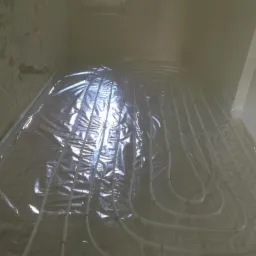 Białe rury ogrzewania podłogowego ułożone spiralnie na folii z siatką w surowym wnętrzu łazienki przed wylewką, widoczne ślady po instalacjach na ścianach.