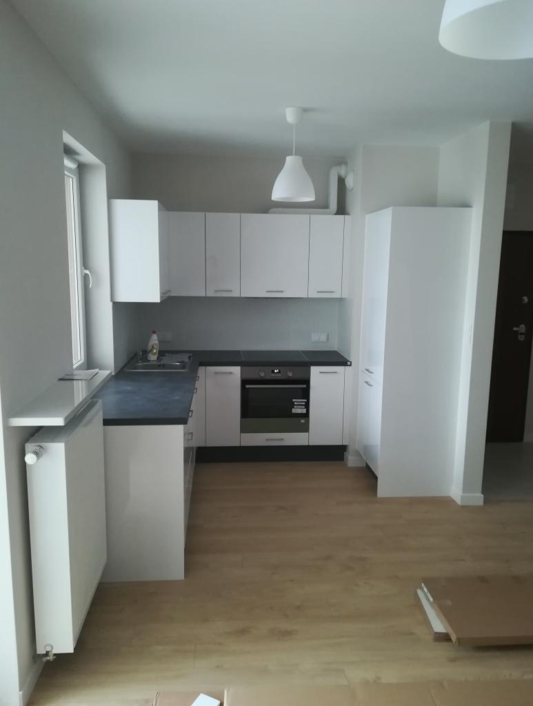 Jasna, minimalistyczna kuchnia po remoncie z białymi szafkami, czarnym blatem, drewnopodobną podłogą i grzejnikiem pod oknem, widoczne kartony po remoncie na podłodze.