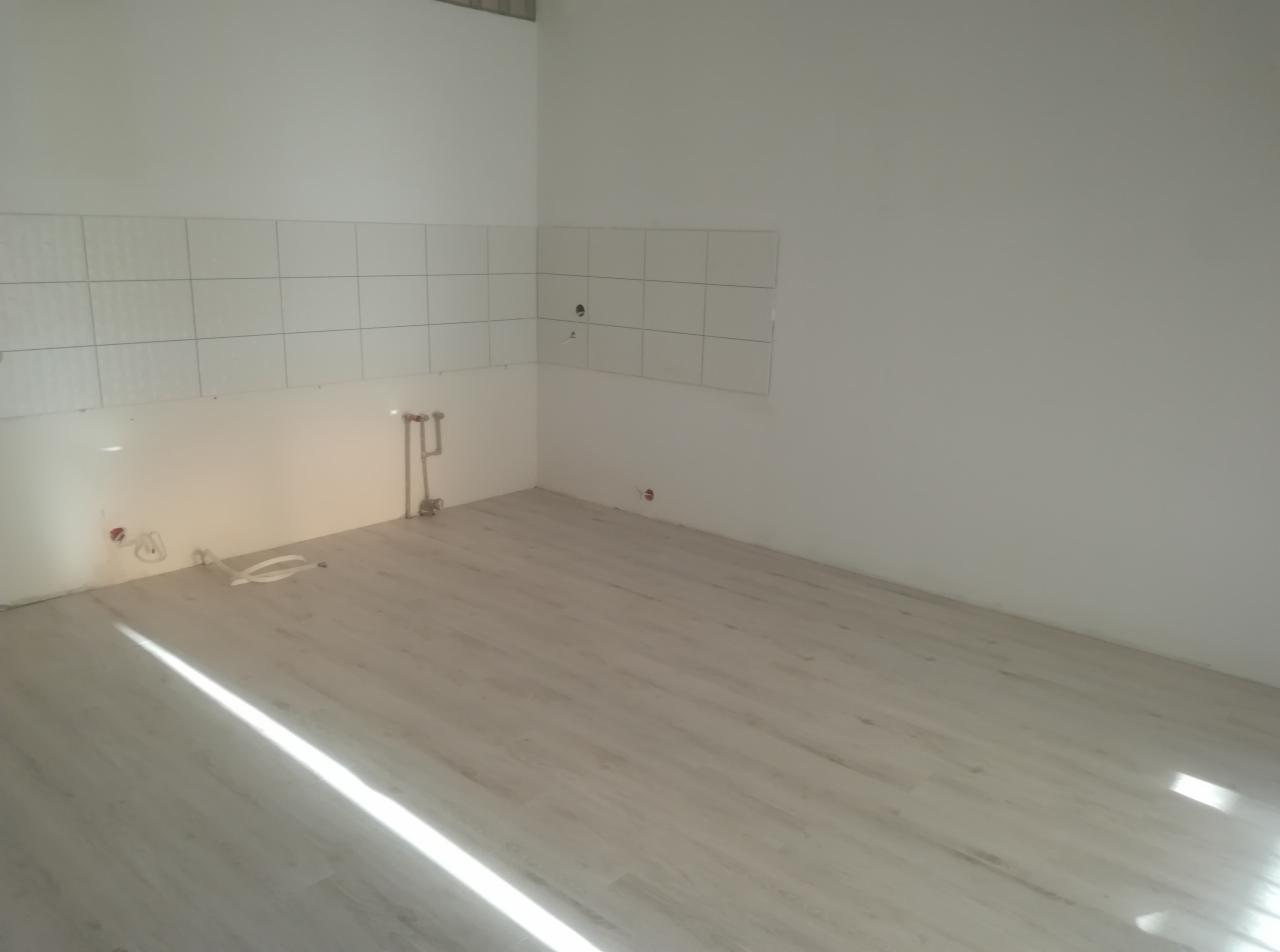 Pusta kuchnia po remoncie, z ułożonymi panelami podłogowymi w odcieniach szarości i częściowo wyłożonymi białymi płytkami ścianami, widoczne wystające rury instalacji wodnej.