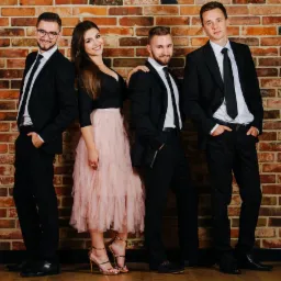 Czteroosobowy zespół muzyczny pozuje na tle ceglanej ściany. Trzech mężczyzn w czarnych garniturach i kobieta w czarnej bluzce i różowej spódnicy tiulowej.