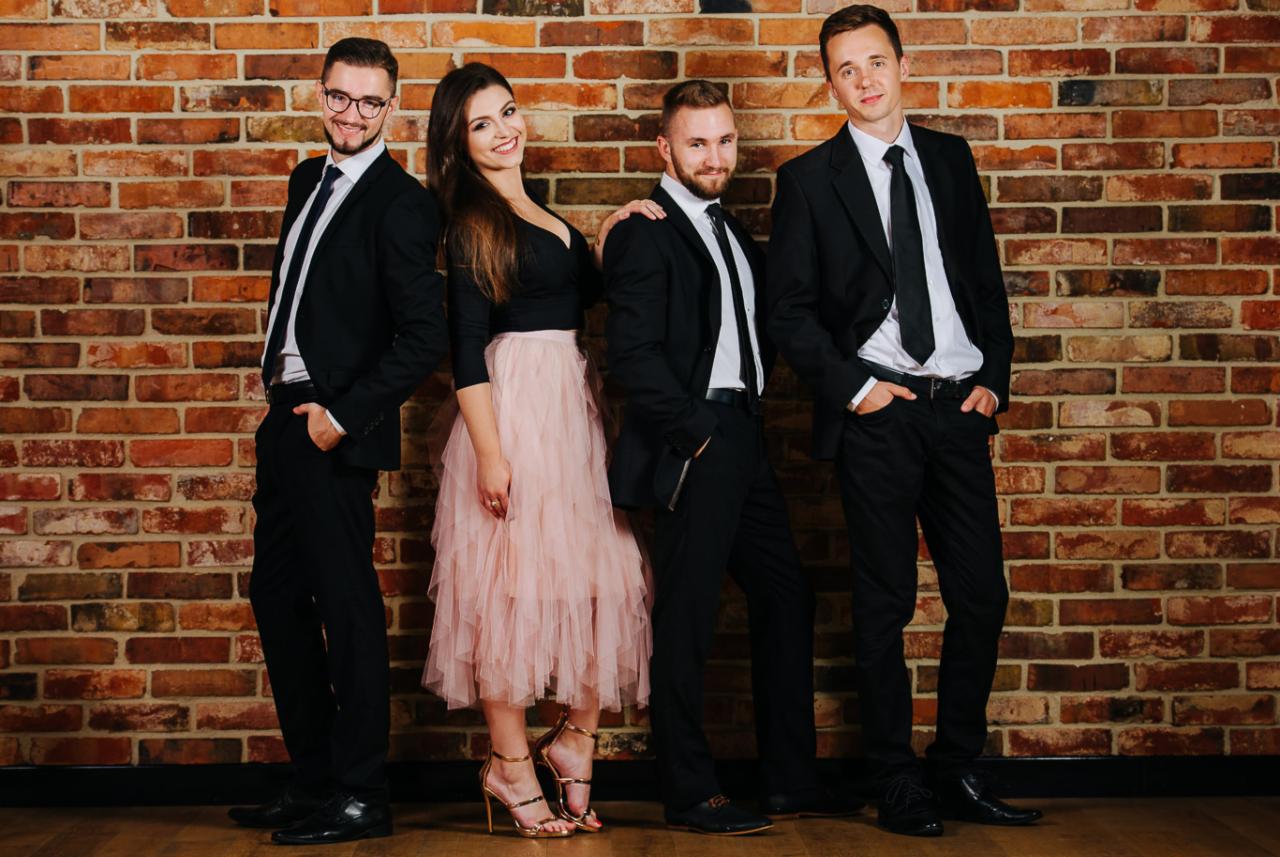 Czteroosobowy zespół muzyczny pozuje na tle ceglanej ściany. Trzech mężczyzn w czarnych garniturach i kobieta w czarnej bluzce i różowej spódnicy tiulowej.