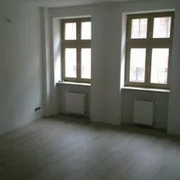 Pusty pokój po remoncie z drewnianą podłogą, dwoma oknami z drewnianymi ramami i białymi ścianami, widoczne grzejniki pod oknami i wisząca żarówka.