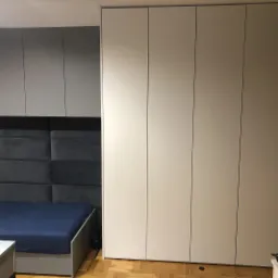 Biała, wysoka szafa wnękowa z przesuwnymi drzwiami, dopasowana do zabudowy sypialni z łóżkiem i panelem tapicerowanym za wezgłowiem.