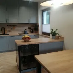 Szara kuchnia z drewnianym blatem, wyspą z przeszkloną witryną na wino i owocami w misie na blacie.